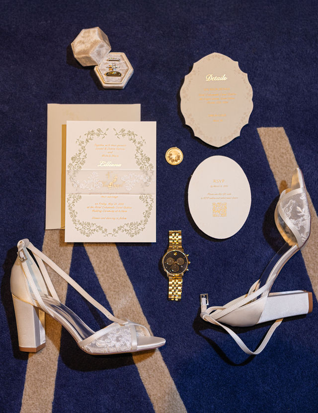 The couple’s wedding stationery, rings and shoes arranged on navy fabric
