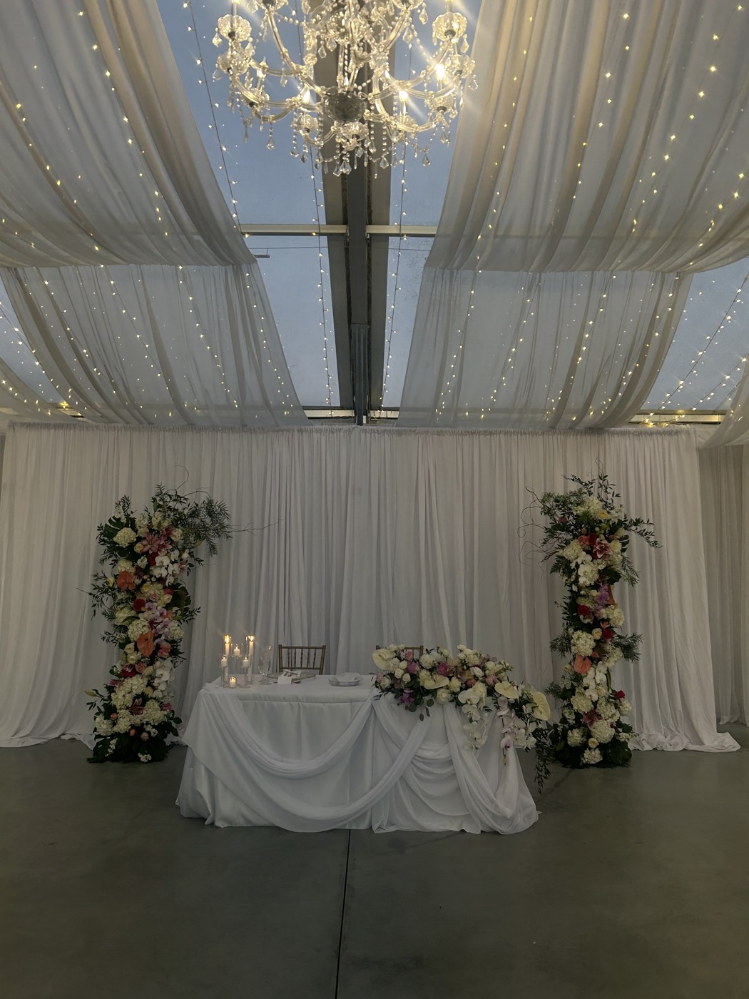 Ivory draping, twinkle lights, a chandelier, and floral columns surrounding a sweetheart table